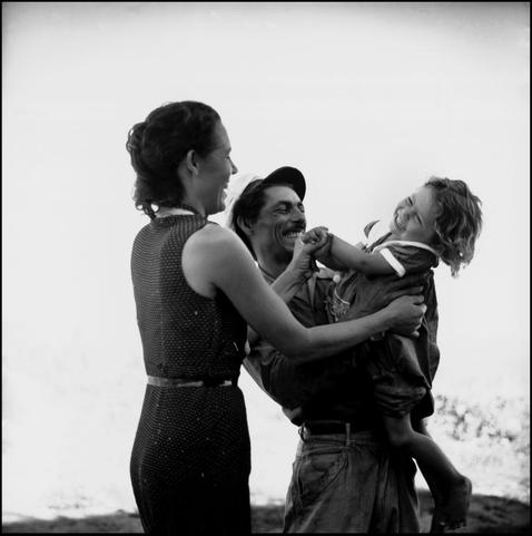 Black and white photo of a white man and woman smiling and holding up and spinning around a small child, against a big empty sky