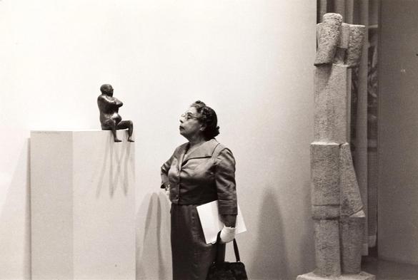Black and white photo of a bespectacled woman in a suit holding a bag and sheet of papers, standing with her hand on hip looking at a small sculpture of a seated man, in a gallery at the Museum of Modern Art in NY