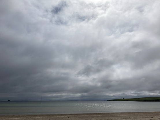 A glimpse of blue sky and sun in a grey cloudy sky. Light reflected in the bay beyond the beach 