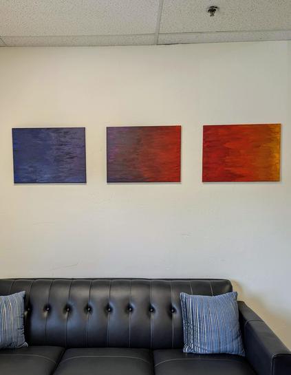 Photo of a couch in an office with three horizonal unframed canvases spaced out with color progressing from blue through purple to red to orange to a dash of yellow.