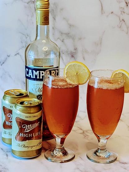 Bottle of campari, 2 open cans of Miller High Life and 2 glasses with a reddish orange drink with a lemon wheel on rim