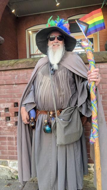 middle aged man with long beard and gandalf style outfit with hat & staff done up in pride colors