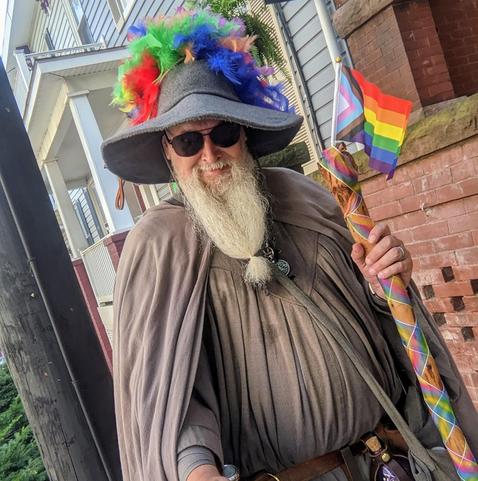 middle aged man with long beard and gandalf style outfit with hat & staff done up in pride colors