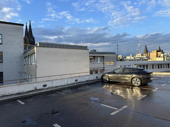 Foto zeigt ein batterieelektrisches KFZ der Marke Kia auf einem Parkhausdach in der Kölner Innenstadt. Auto ist schwarz, Himmel leicht bewölkt, Parkdeck vom Regen nass, hinter irgendwelchen Fassaden sind die Kirchturmspitzen der örtlichen katholischen Kirche zu erkennen. 