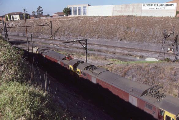 A view looking down from an embankment at the side of a railway line overlooking a pair of predominantly Indian Red liveried boxcab electric locomotives.  The lower half of the loco sides are in the shadow of the embankment.  In the background another couple of tracks are visible and another tall embankment is in the distance with a slab-sided industrial building on the top.