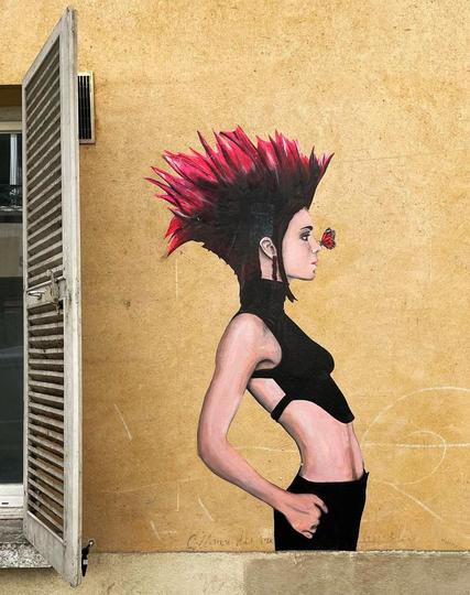 Streetartwall. The charming paste-up of a punk girl with a butterfly was stuck on a yellow exterior wall next to a window. The slender young woman is pictured up to her waist and sideways. She has spiky red hair and is wearing a black crop top and black pants. The paste-up is made cute by the little brown butterfly that has landed directly on her nose.
