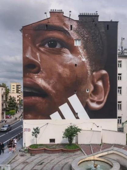 Streetartwall. Half of the face of black footballer Jude Bellingham has been painted on the outside wall of a four-storey building. He is looking upwards. The mural is photo-realistic and was commissioned by a well-known sportswear manufacturer. The three white stripes can therefore be seen.