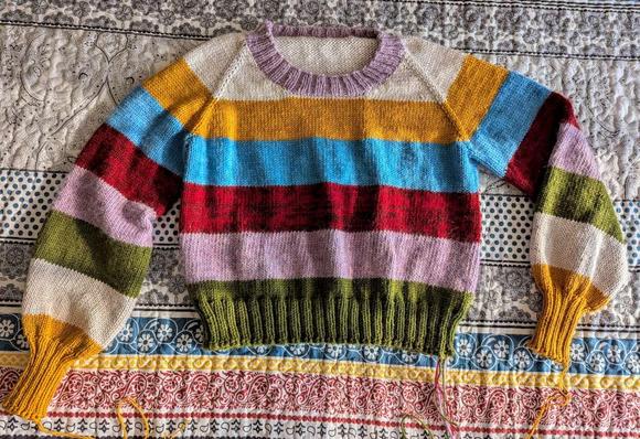 A short pullover sweater with long puffed sleeves, laying on a bedspread that is mostly black and white. It has 6 wide stripes: white, orange-yellow, blue, red, pink, and olive green. The collar is pink, the cuffs are orange-yellow, and the waistband is green.