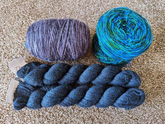 Four skeins of yarn on brown carpet. One is shades of gray and sparkly, one is alternating blue and green, and two are dark navy blue.