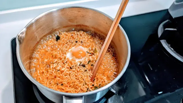 Korean ramyeon instant noodles being cooked with an egg in a pot. The noodles are in a bright red-orange broth with pieces of kelp and scallions.