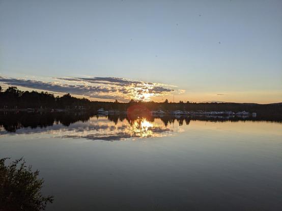 Magnifique non-coucher de soleil (le soleil descend vers l'horizon nord mais ne se couche pas réellement à cette période ici) au-dessus du lac Inari. On aperçoit un petit port de plaisance au centre au fond, la berge est bordée d'arbres. Le ciel est bleu avec seulement quelques nuages qui cachent le soleil, dont la lumière se reflète dans le lac.
