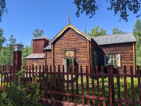 L'église en bois du lac Pielpajärvi près d'Inari, entourée d'une clôture en bois et d'arbres. Le ciel est bleu.