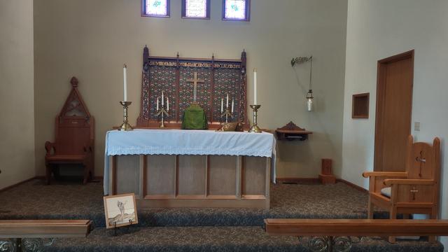 The altar at my Episcopal Church (St. Timothy's). Sitting in front of it is a small icon depicting the crucifixion. The altar itself is adorned by a white cloth, two candles sit at either end. An open Bible sits in a brass book holder. In the back are two three-candle holders sitting on either side of a covered holy oil vessel. Situated directly above is a plain brass cruciform. To the left of the altar is a chair, to the right is a small, wall mounted table, the holy Spirit candle hanging beside it, the crucifer mount, another chair sits to the left of the door to the room where the Holy Communion is prepared.