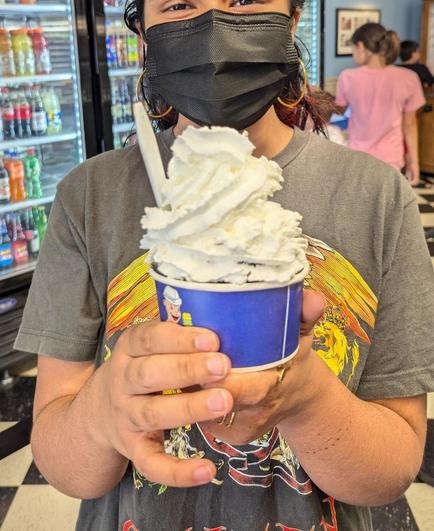 Kid holding a paper cup (filled with a scoop of chocolate ice cream, not visible, with) a tall heap of whipped cream on top