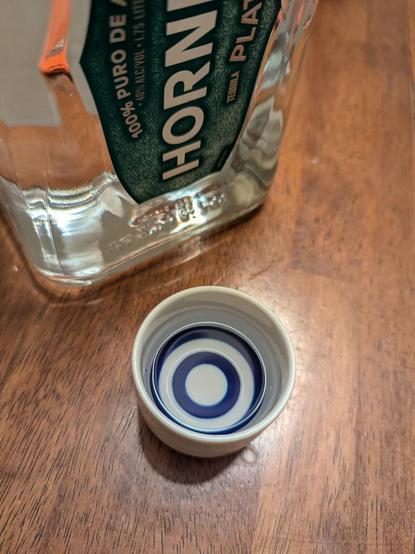 A bottle of Blanco tequila and a small white porcelain sake cup with blue rings at the bottom