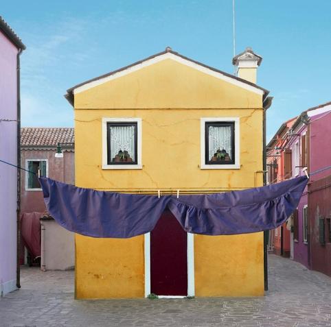 Photography and art. A color photo of a yellow house with a mustache. A small one-story house with a brown wooden door and two windows stands on a village square. White curtains are draped in a V-shape in the small windows so that a plant decoration can be seen in the middle. A washing line with purple bed linen is stretched across the front of the house. All this together makes for a very funny photo. The purple bedding in front of the house looks like a giant moustache, the door looks like an open mouth and the windows look like eyes. A funny and astonished looking house with a moustache.