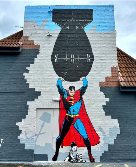 Streetartwall. A large mural of Superman was spray-painted on a one-story exterior wall between the roofs of a rooftop parking lot. Superman stands with his amen facing upwards, holding up a giant bomb. He is looking down as a small baby sits between his legs on the ground playing with his toy cars. (Superman is depicted in blue and red, the child in gray/black/white).
Info: The artist says:  
