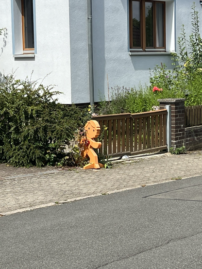 A small orange figure, designed to raise awareness that children are playing in the neighbourhood, and you should drive carefully, peaks outpeaks out from behind a bush. His orange colour and haircut reminds me of Trump.