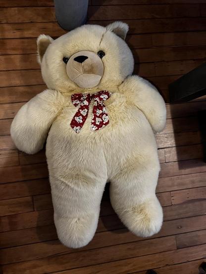 The image features a large, plush teddy bear sitting on a wooden floor. The teddy bear is light beige in color with darker beige accents on its snout and paw pads. 