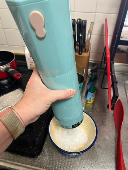 A hand holds a cylindrical pale blue shaved ice maker over a bowl containing ground up ice. 