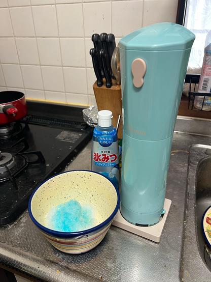 The bowl of shaved ice beside the shaved ice maker sits in the kitchen beside the sink. The shaved ice is now blue from an added flavour bottle that sits behind it. 