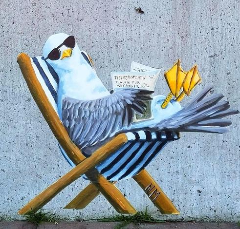 Streetartwall. A mural with a chilling seagull was painted on a gray concrete wall. The seagull with sunglasses sits smiling in a deckchair with a german book: 