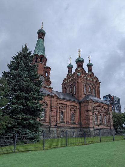 L'église orthodoxe de Tampere, en brique rouge et toiture verte. Au premier plan à gauche, un gros sapin. Le ciel est gris.