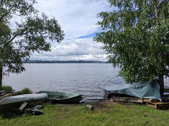 Vue sur l'un des deux lacs de Tampere, côté sud. Au premier plan des barques sur la berge et des arbres de part et d'autre. Au loin l'autre rive, sur laquelle on distingue des villes et des forêts. Le ciel est nuageux avec quelques taches de bleu.
