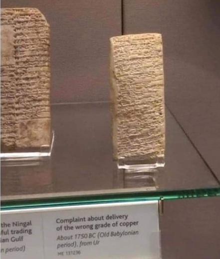 A clay block sits on a glass museum shelf. On it is scribed cuneiform writing. Below the shelf a note explains the block is a note complaining about a delivery of copper being the wrong grade. The date of the block is stated as 