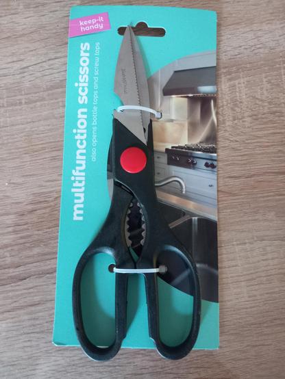 A pair of household scissors is shown in their original packaging, held firmly by two cable ties 
