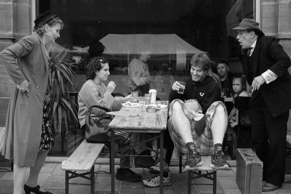 Two people are having a relaxed outdoor Sunday lunch at a Glasgow cafe. Two performers have intruded in a comic fashion, while people inside the cafe look out in confusion