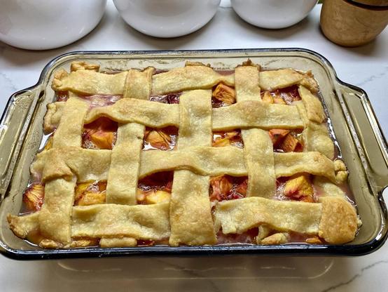 Photo of a lattice topped fruit cobbler in a Pyrex baking dish. The fruit inside is peaches. You can see them peaking out between the holes in the lattice top.
