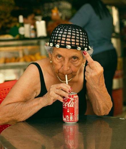 Photography. A color photo.of an old lady drinking a can of Coca Cola and giving the photographer the middle finger. The woman is wearing a large-mesh, crocheted headdress with a wreath of small shells and a black top.  She is leaning on the tabletop with her elbows and has a straw in her mouth with which she is drinking from a can of Coke. She clearly shows how she feels about being photographed and gives the photographer, and therefore us, the middle finger. (A café counter with a woman can be seen blurred in the background.)