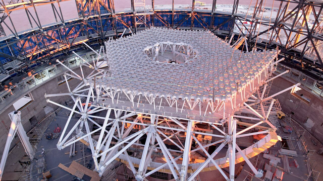 The image shows a partially-constructed white hexagonal lattice structure with a honeycomb-like design, in the interior of a metallic dome structure with some blue cladding on the exterior. In the centre of the lattice structure, there is a hexagonal hole in the structure.
