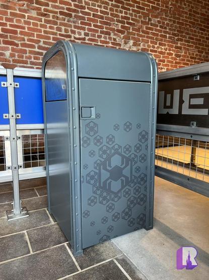 A silver trash can machine with a hexagonal and spider motif set against a brick wall background. There is a metal barricade and a sign with partial text visible to the right.