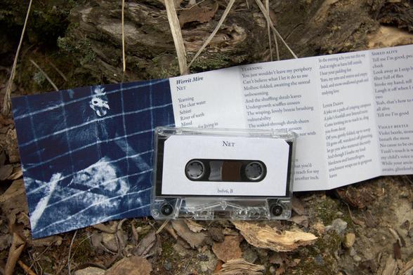 Promotional photograph of a music cassette tape along with its accompanying insert. The tape and insert are positioned outside on the ground in woods.