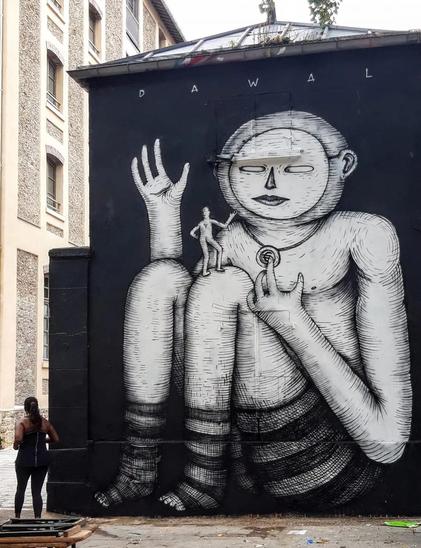 Streetartwall. The mural of a giant was painted in black and white on a small one-story house. On a black background sits a white giant with a round, friendly face, dressed in striped shorts and stockings and wearing a necklace. With one of his four fingers he touches the pendant, which shows a euro currency symbol, and waves with the other hand. A small man stands on his knees and also waves to him. (The windows and doors have been painted over and are barely visible).