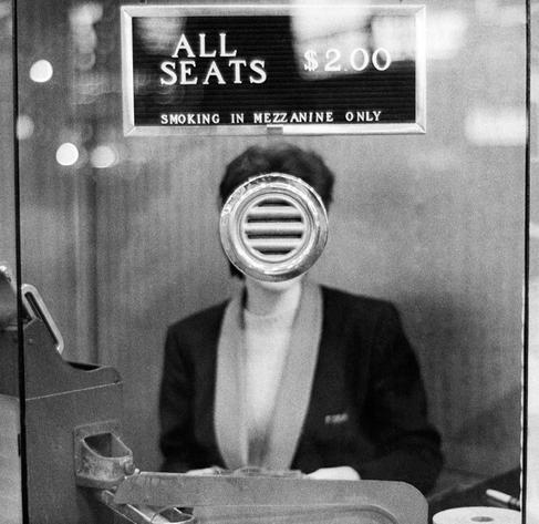Photography. A black and white photo of a cashier in the entrance area of a theater. The cashier's face, standing in the center of the image, is completely obscured by the small metal intercom window through which one would speak to her, giving the lady in the black costume a surreal faceless appearance. Above her hangs a sign that reads, 
