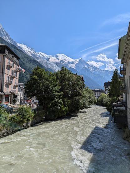 A view of the mont blanc mountain range from chamonix. A rushing canal full of cold glacial water is in the foreground, in between buildings