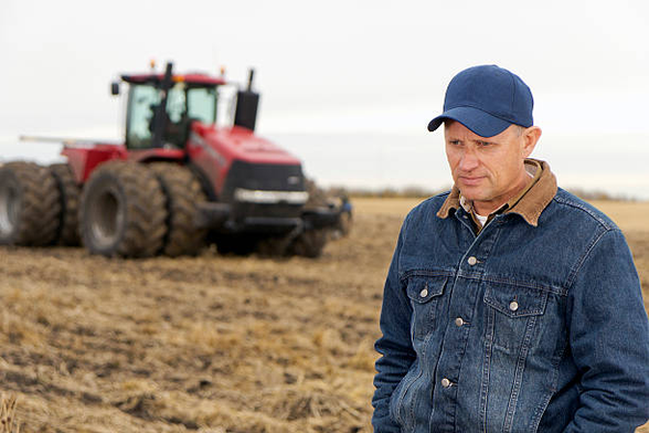 Photo of a farmer standing in his field near a tractor, looking down and looking sad.