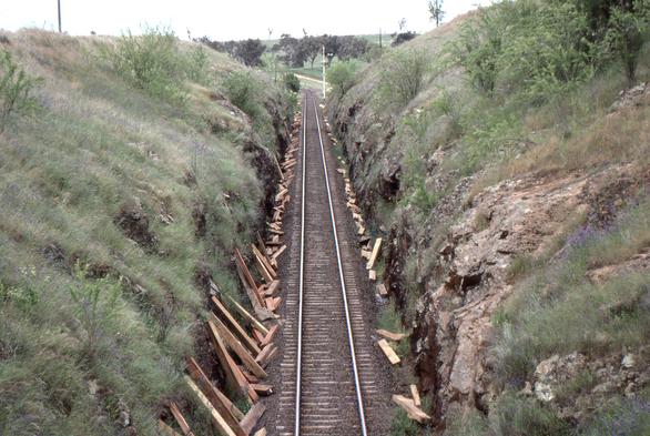 A single track railway line in a moderately deep rock and grass sided cutting.  New wooden sleepers are lying against the cutting edges along the length of the track in readiness for re-sleepering.