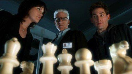 In a scene from CSI, Jorja Fox, Ted Danson and Eric Szmanda look down at a chess set that looms in the extreme foreground.