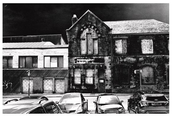 Adjusted black and white dark film photograph showing abandoned derelict buildings behind a line of parked cars.