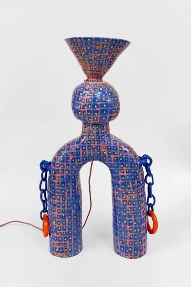 Abstract ceramic sculpture standing on two rounded legs with a funnel shape at the top over a rounded center, and two chains emerging on either side. The surface is covered in a loose grid in blue and pink with white circles in each square's center