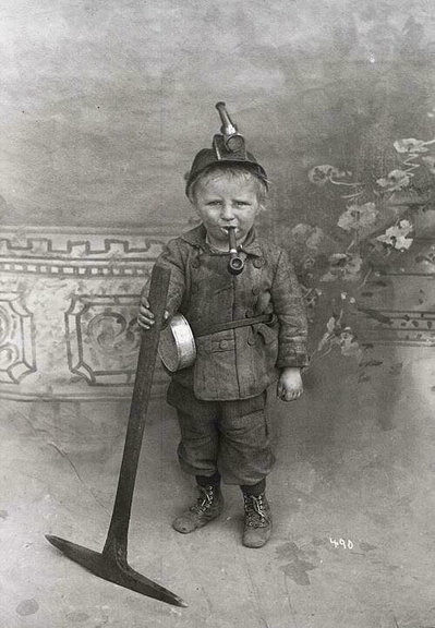 A staged picture of a child miner probably from the 1900s.  The child has a pipe in his mouth, is wearing a mining hat with a light and is holding a full size pick axe.

