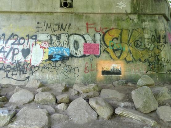 Photograph of a concrete wall bearing a variety of overlapping graffiti.  Most prominent are three relatively small rectangular pieces, in bright blue, pink, and orange.
