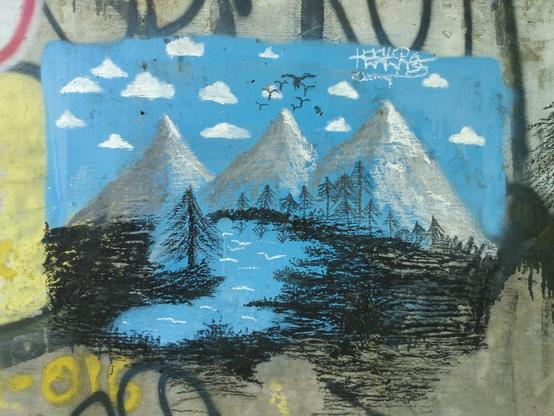 Close-up photograph of the primarily blue piece.  From foreground to background, it depicts a tree by a lake, a row of several trees, three snow-capped mountains, and a sky filled with clouds and a flock of birds.  It is signed 