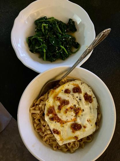 Two white bowls sit on a dark table. They are positioned next to each other. One contains a portion of spinach that is dark green and flecked with toasted sesame seeds. The other bowl is slightly larger and contains wavy noodles coated in a brown sauce with a fried egg on top. The egg is dotted with bright red chili garlic crunch. A fork is sticking out of the bowl containing the noodles.