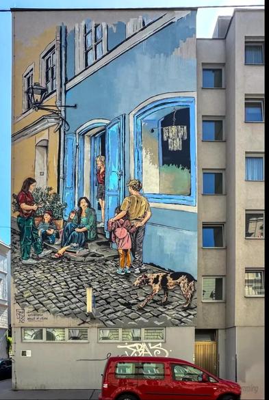 Streetartwall. A large mural (15.6 x 7.9 m) depicting a street scene was painted on the outside wall of a five-storey building. A family with neighbors in front of a blue store was painted in wonderful detail like an artistic pastel drawing. A blonde young woman in shorts stands in the doorway, a long-haired woman in a blue skirt sits on the steps in front of green plants, two little girls stand around them and a young woman stands in front of them playing the ukulele. A coat hanger with an item of clothing hangs in the shop window. A blond boy stands with his back to us, arm in arm with a smaller girl. And in the foreground of the picture, a brown dog runs across the cobblestones. What a wonderful narrative in picture form.
Info: This interpretation is based on the painting 
