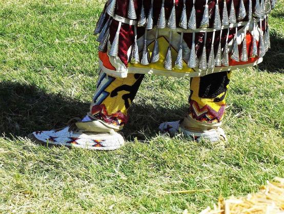 Lower legs wearing traditional Native American regalia with colorful beadwork, mocassins  and jingles, standing on grass.